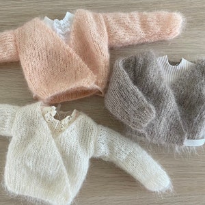 Peut inclure: Trois cardigans en tricot pour bébé en rose, gris et blanc. Les cardigans sont faits d'un fil doux et duveteux et ont un style enveloppant.