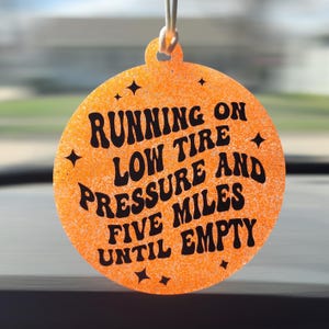 May include: Orange, circular car air freshener with black text that reads "RUNNING ON LOW TIRE PRESSURE AND FIVE MILES UNTIL EMPTY." The air freshener has a glittery texture and four black star accents.