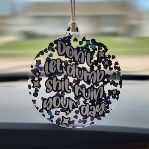 May include: A clear, round car charm with the text "Don't let dumb shit ruin your day" in black script. The charm is filled with iridescent heart and star-shaped glitter, and hangs from a white string.