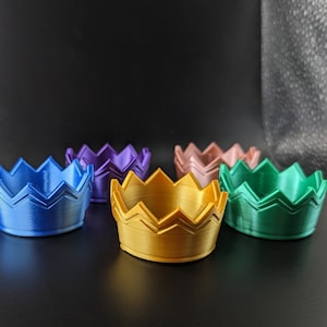 May include: Five small, decorative crowns in different colors. The crowns are made of plastic and have a zig-zag pattern. The colors are blue, purple, pink, gold, and green.