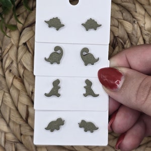 May include: Four pairs of green dinosaur-shaped stud earrings on a white card. The earrings are arranged in four rows, with two earrings in each row. The earrings are made of a textured material that resembles wood.