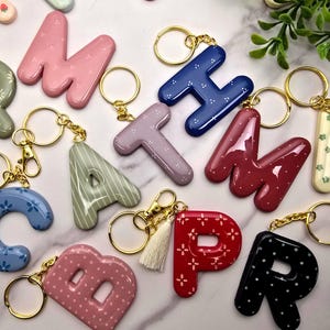 May include: A collection of letter-shaped keychains in various colors including pink, blue, green, red, and black, featuring patterns like dots, stripes, and flowers. Each keychain has a gold-toned metal ring and clasp, with one red letter featuring a small tassel.
