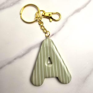 May include: A sage green letter A keyring featuring a glossy finish and thin vertical white stripes. The letter is attached to a gold-toned metal split ring and a lobster claw clasp by a short gold-toned chain link connector.