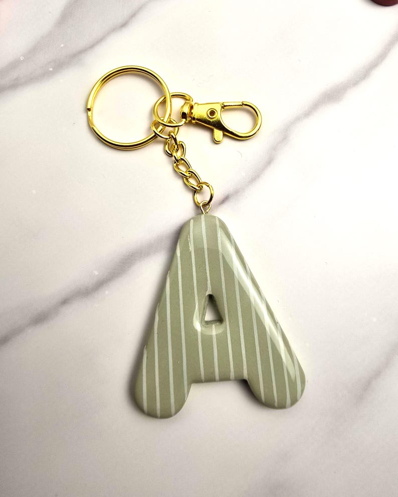 May include: A sage green letter A keyring featuring a glossy finish and thin vertical white stripes. The letter is attached to a gold-toned metal split ring and a lobster claw clasp by a short gold-toned chain link connector.