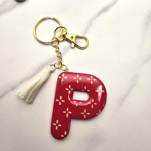 May include: A glossy red letter P keyring featuring a white cross-stitch pattern. The letter is attached to a gold-toned metal split ring and lobster clasp, accompanied by a small, off-white fabric tassel hanging from the gold chain link.