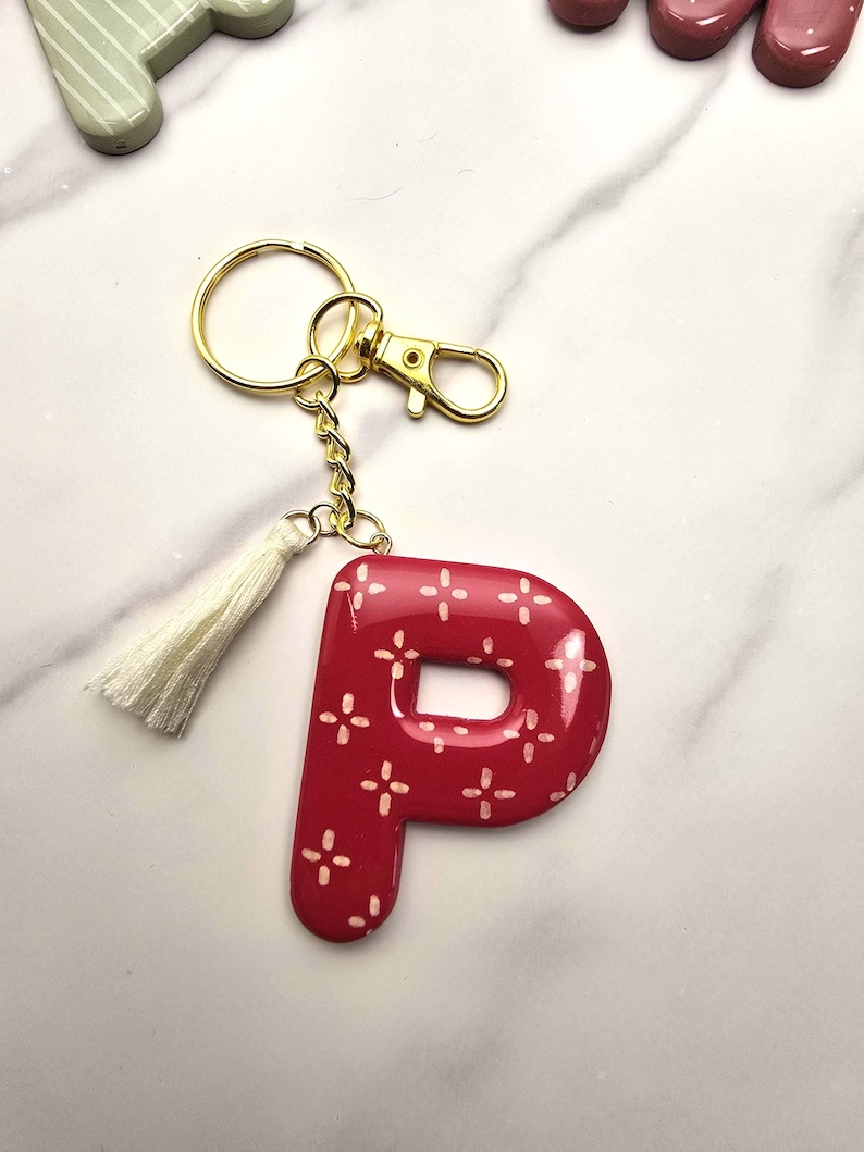 May include: A glossy red letter P keyring featuring a white cross-stitch pattern. The letter is attached to a gold-toned metal split ring and lobster clasp, accompanied by a small, off-white fabric tassel hanging from the gold chain link.