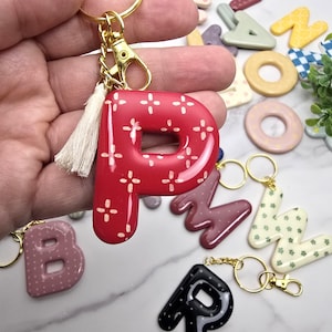 May include: A glossy red letter P keyring featuring a white cross-stitch pattern, attached to a gold-toned metal ring and lobster clasp with a small cream-coloured fabric tassel. The letter has a smooth, rounded finish and a shiny surface texture.