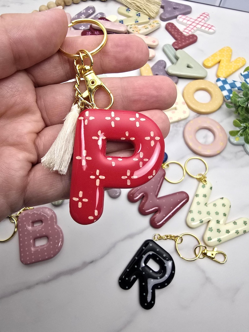 May include: A glossy red letter P keyring featuring a white cross-stitch pattern, attached to a gold-toned metal ring and lobster clasp with a small cream-coloured fabric tassel. The letter has a smooth, rounded finish and a shiny surface texture.