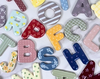 May include: A collection of colourful, glossy alphabet letters featuring various patterns including floral, polka dot, striped, and fruit designs. Each letter is distinct in colour and print, scattered across the frame in a random arrangement.