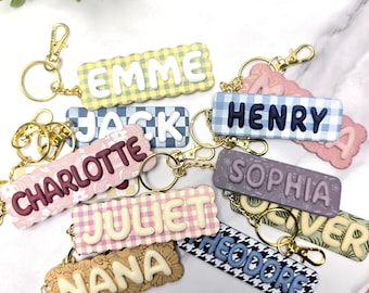 May include: A collection of rectangular keychains with scalloped edges, each featuring a raised name in a contrasting colour. The keychains display various patterns including gingham, floral, and houndstooth, and are attached to gold-toned metal lobster claw clasps and rings.