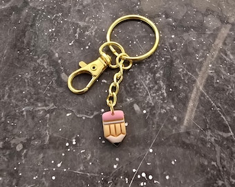 May include: A gold-coloured keyring with a pencil charm. The keyring has a lobster clasp, a small chain, and a pencil-shaped charm with pink, tan, and white details. The pencil charm is a miniature version of a classic pencil.