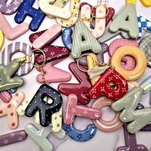 May include: A collection of glossy, three-dimensional letter keychains in various colours and patterns, including floral, gingham, and polka dot designs. Several letters feature gold-toned metal key rings, with one letter attached to a small beige tassel.