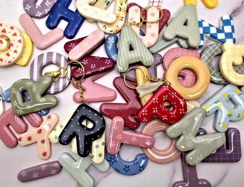 May include: A collection of glossy, three-dimensional letter keychains in various colours and patterns, including floral, gingham, and polka dot designs. Several letters feature gold-toned metal key rings, with one letter attached to a small beige tassel.