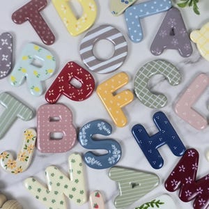 May include: A collection of colourful alphabet letters featuring various patterns including floral, polka dot, striped, and checkered designs. The letters are rendered in shades of yellow, blue, pink, red, green, and brown with a smooth finish.