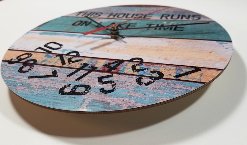 Puede incluir: Un reloj redondo de madera con un acabado pintado desgastado. La esfera del reloj tiene n&uacute;meros negros y el texto "This house runs on lake time".