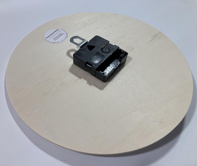 Puede incluir: La parte posterior de un reloj redondo blanco con un mecanismo de reloj negro adjunto. El mecanismo del reloj tiene una peque&ntilde;a pegatina que dice "Crazy Clock Shop".