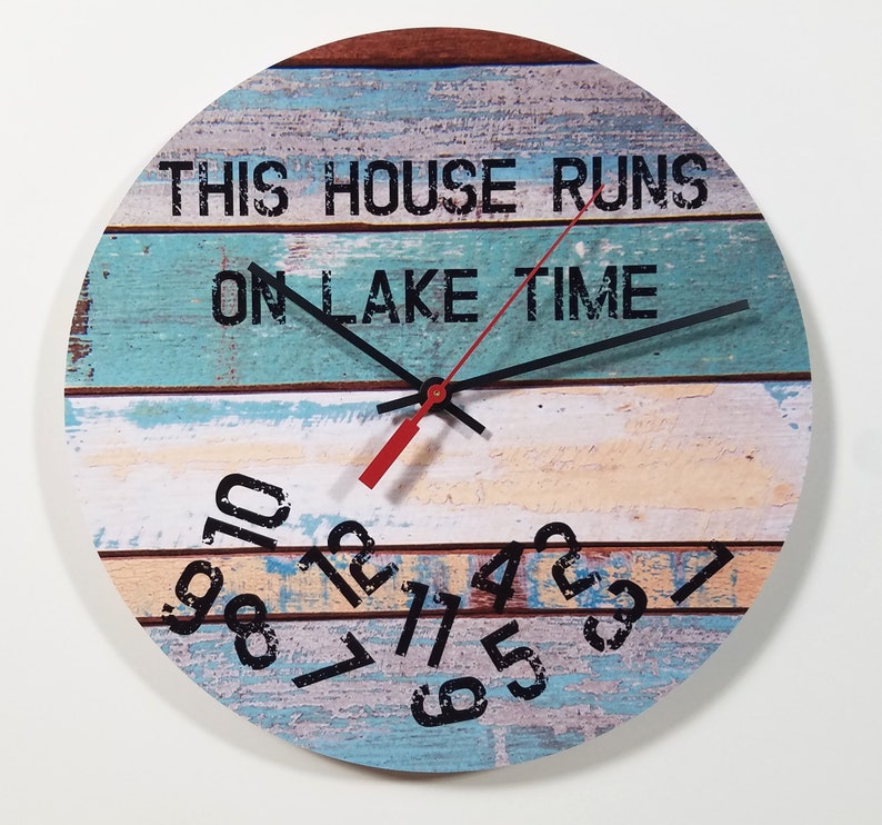 Puede incluir: Un reloj redondo de madera con un dise&ntilde;o a rayas azul, blanco y marr&oacute;n. La esfera del reloj tiene n&uacute;meros y manecillas negros, y el texto "This house runs on lake time".