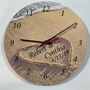 May include: A round wooden clock with a beach scene background. The clock face has Roman numerals and a heart drawn in the sand with the text "Robert & Cynthia 4/12/19".