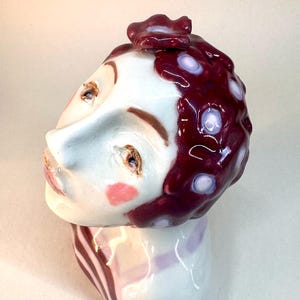 May include: A ceramic bust sculpture featuring a stylized head with a white glazed surface, painted facial features, pink cheek accents, and a dark maroon hair section adorned with light purple circular patterns and a small sculpted flower detail.