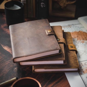 May include: Three brown leather journals with a worn look and a brass closure. The journals are stacked on top of each other on a wooden surface.