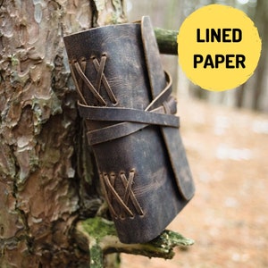 May include: A brown leather-bound journal with a rustic look. The journal is closed and has a leather strap with decorative stitching. The journal is sitting on a tree branch.