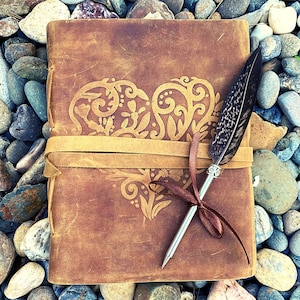 May include: A brown leather journal with a heart-shaped design and a feather pen. The journal has a leather strap and a decorative metal tip on the pen.