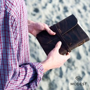 May include: A brown leather journal with a strap closure. The journal is closed and held in the hands of a person.