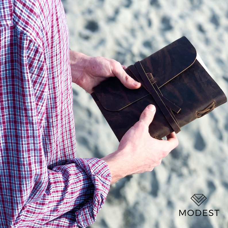May include: A brown leather journal with a strap closure. The journal is closed and held in the hands of a person.