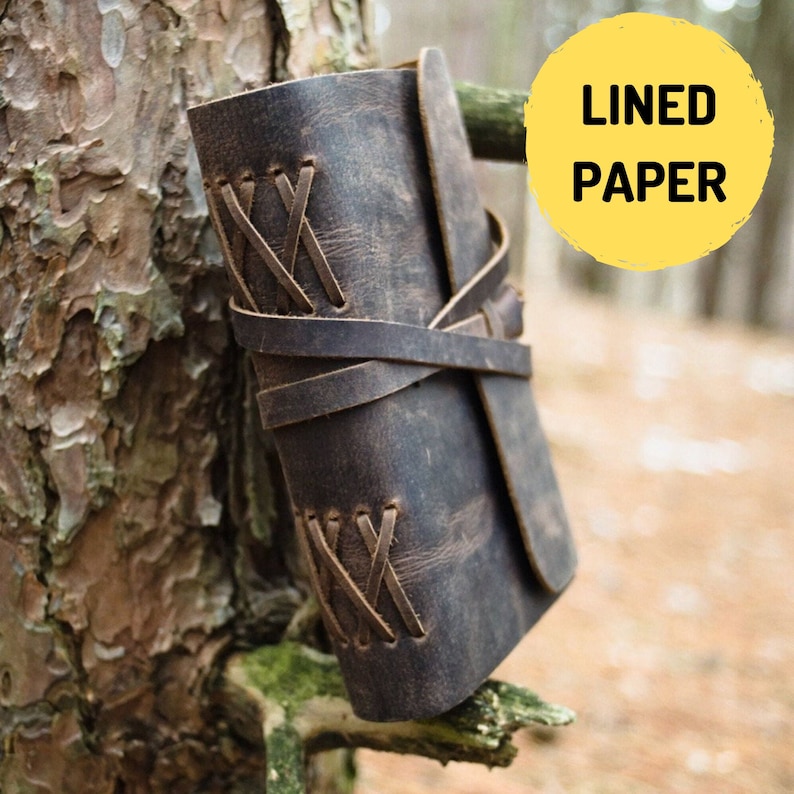 May include: A brown leather-bound journal with a rustic look. The journal is closed and has a leather strap with decorative stitching. The journal is sitting on a tree branch.
