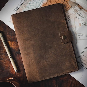May include: A brown leather journal with a flap closure and a brass button. The journal has a distressed look and is perfect for writing or sketching.