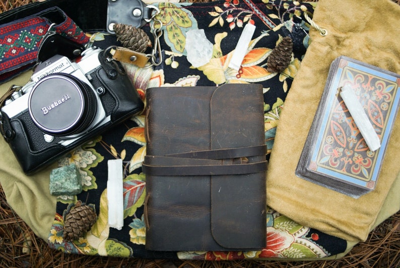May include: A vintage camera, a leather-bound journal, a bag of tarot cards, and various crystals and stones are arranged on a floral fabric background.