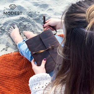 May include: A brown leather journal with a tan leather strap closure. The journal is closed and held by a person sitting on a beach.