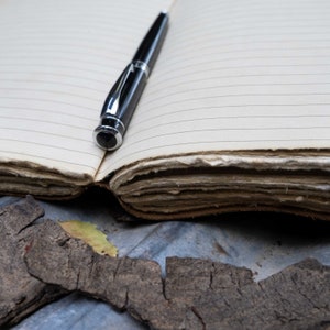 May include: A black pen rests on a blank, lined notebook with aged, worn pages. The notebook is open and the pen is positioned near the center of the page.