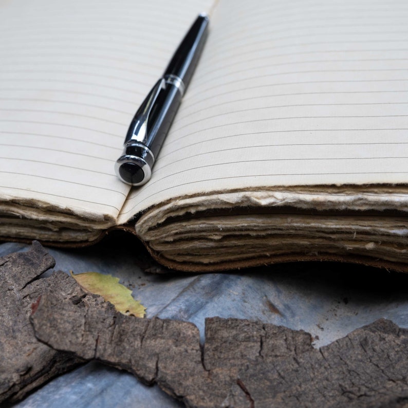 May include: A black pen rests on a blank, lined notebook with aged, worn pages. The notebook is open and the pen is positioned near the center of the page.