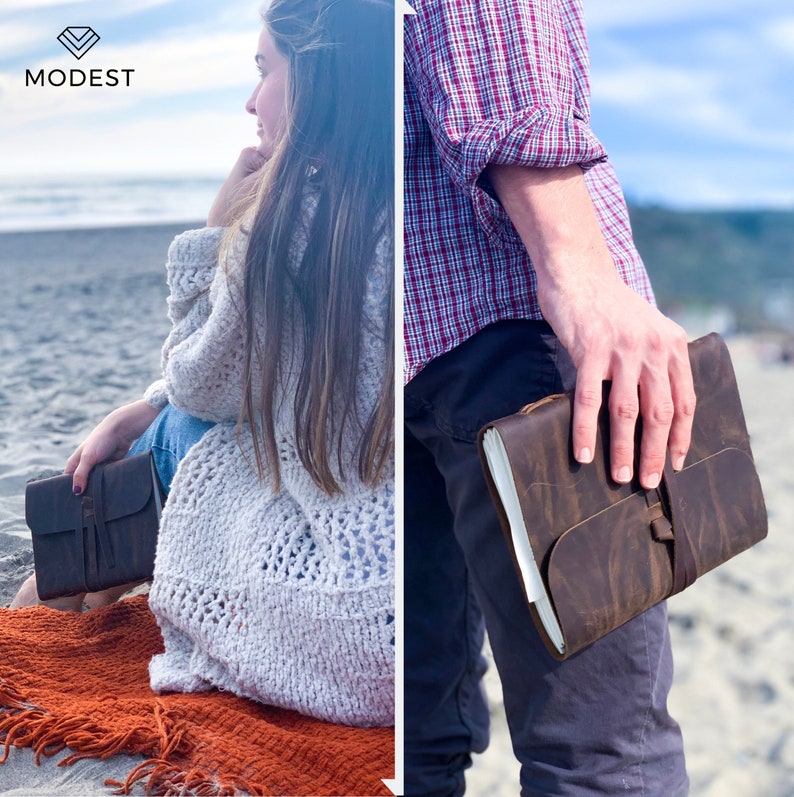 May include: A brown leather journal with a strap closure. The journal is held by a person wearing a plaid shirt and jeans. The journal is on a beach with a person sitting in the background.