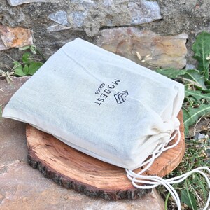 May include: A white drawstring bag with the text "WOODEST GOODS" printed on it. The bag is sitting on a wooden surface.