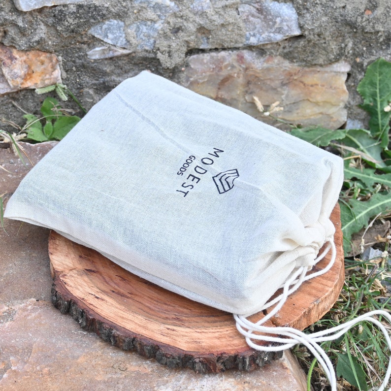 May include: A white drawstring bag with the text "WOODEST GOODS" printed on it. The bag is sitting on a wooden surface.