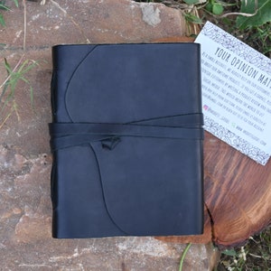 May include: A black leather-bound journal with a strap closure. The journal is sitting on a wooden surface. The text on the card reads "Your Opinion Matters".