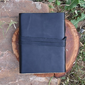 May include: A black leather-bound journal with two stitched straps. The journal is closed and sitting on a wooden surface.