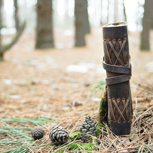May include: A brown leather journal with a criss-cross pattern on the cover. The journal is sitting on a bed of pine needles in a forest setting.