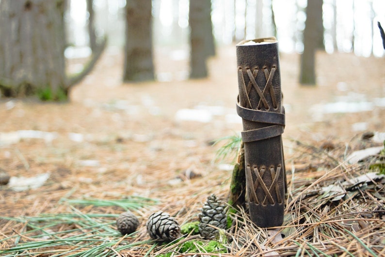 May include: A brown leather journal with a criss-cross pattern on the cover. The journal is sitting on a bed of pine needles in a forest setting.