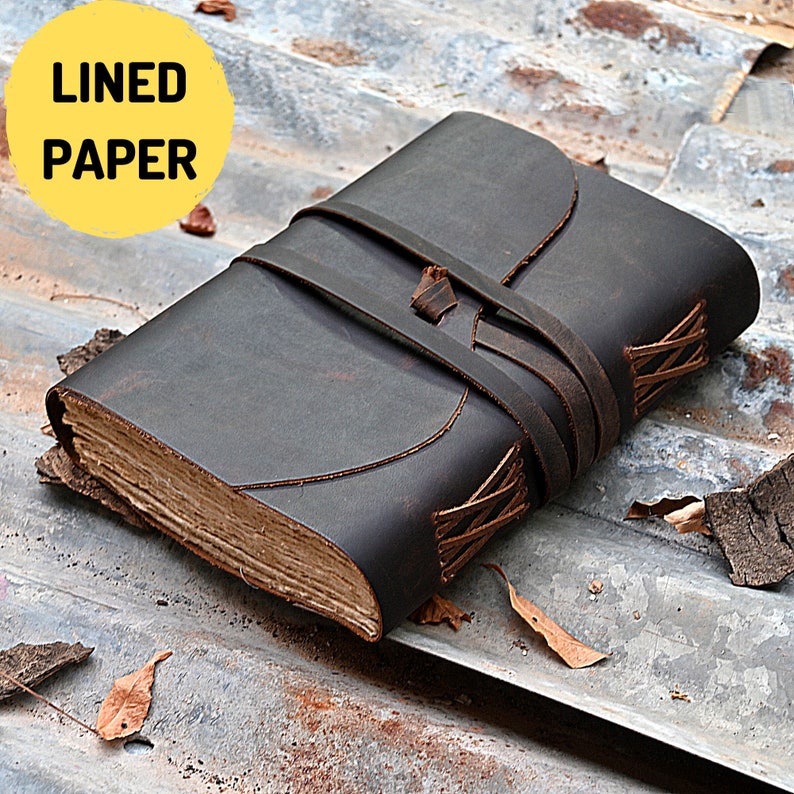 May include: A brown leather-bound journal with a lined paper interior. The journal has a leather strap closure and is closed.