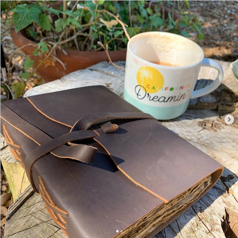 May include: A brown leather journal with a strap closure and decorative stitching. The journal is sitting on a wooden surface next to a white mug with the text "California Dreamin'" on it.