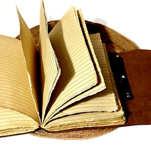 May include: A brown leather-bound journal with lined pages is open and fanned out. The journal has a metal clasp and is resting on a woven mat.