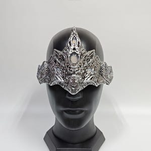 Może przedstawiać: Srebrna korona o skomplikowanym wzorze i białych kamieniach szlachetnych. Korona jest noszona na czarnej głowie manekina.