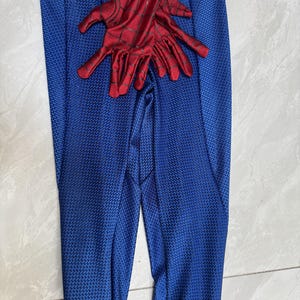Puede incluir: Un mono de cuerpo entero rojo y azul con un patr&oacute;n de panal texturizado en las secciones azules y un dise&ntilde;o de telara&ntilde;a negra en las partes rojas. La prenda incluye guantes rojos a juego con telara&ntilde;a negra sobre el &aacute;rea del torso.