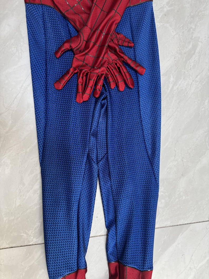 Puede incluir: Un mono de cuerpo entero rojo y azul con un patr&oacute;n de panal texturizado en las secciones azules y un dise&ntilde;o de telara&ntilde;a negra en las partes rojas. La prenda incluye guantes rojos a juego con telara&ntilde;a negra sobre el &aacute;rea del torso.