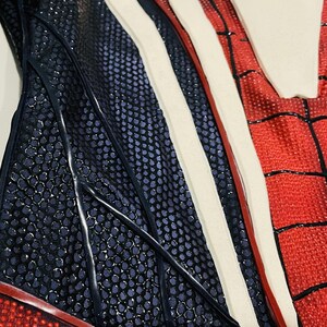 May include: Close-up of a red, white, and blue costume with a textured pattern. The costume features a web-like design in black on the red sections, with white and blue accents.