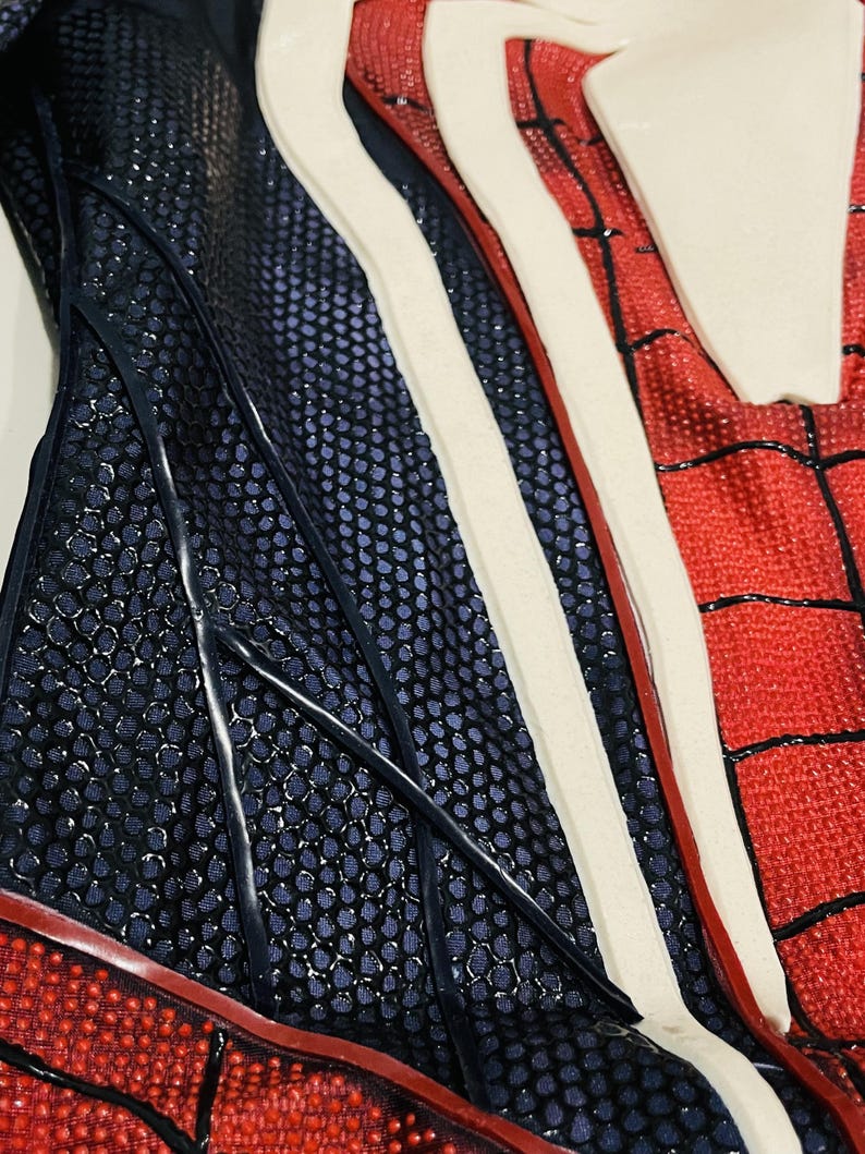 May include: Close-up of a red, white, and blue costume with a textured pattern. The costume features a web-like design in black on the red sections, with white and blue accents.