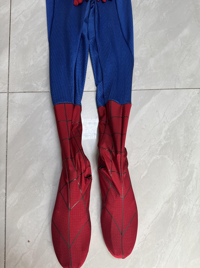 Puede incluir: Un disfraz de cuerpo completo que presenta tela azul con un patr&oacute;n de panal texturizado en la secci&oacute;n superior y tela roja con un estampado de telara&ntilde;a negra en las secciones de la parte inferior de la pierna y los pies, con calzado integrado.